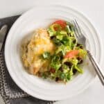 Golden baked dish topped with melted cheese, served alongside a fresh garden salad with tomatoes, black beans, and cilantro on a white plate.